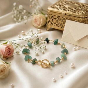 May include: A bracelet featuring irregular, tumbled green stone beads linked by gold-toned wire, secured with a circular toggle clasp. The piece is arranged on a soft, draped fabric surface alongside small white flowers and scattered pearls.