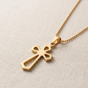 Puede incluir: Un colgante de cruz de metal en tono dorado suspendido de una delicada cadena de bolas a juego. La cruz presenta un diseño hueco con bordes lisos y pulidos, con una silueta sencilla y limpia que se conecta a la cadena mediante una argolla.