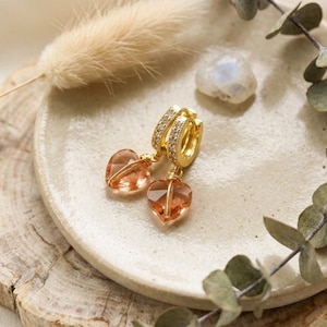 Puede incluir: Un par de pendientes de aro dorados con una hilera de pequeñas piedras transparentes en el borde exterior y un dije colgante de piedra preciosa en forma de corazón de color melocotón. Los pendientes tienen cierre de bisagra y acabado metálico.