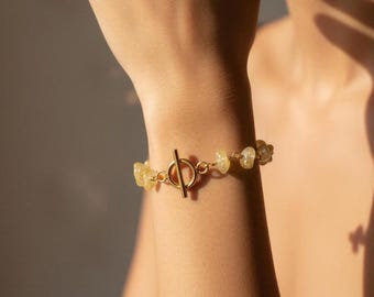 Può includere: Un bracciale a catena color oro con una chiusura a T centrale e piccole pietre traslucide di colore giallo pallido e di forma irregolare intervallate lungo le maglie. Il bracciale è fissato con un meccanismo di chiusura a barra e anello.