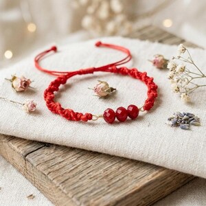 Puede incluir: Una pulsera de cordón rojo con un diseño trenzado y tres cuentas rojas facetadas en el centro. La pulsera incluye cordones rojos finos y ajustables que se extienden desde la sección trenzada principal, permitiendo un ajuste flexible.