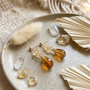 Puede incluir: Un par de pendientes colgantes dorados con cuentas de lágrima facetadas de color ámbar y pequeños detalles de cristal transparente. Los pendientes se muestran junto a varios fragmentos de piedras preciosas sueltas, en bruto y pulidas.