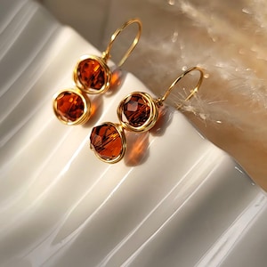 May include: A pair of dangle earrings featuring two faceted amber-colored crystals wrapped in gold-toned wire. Each earring is suspended from a gold-toned hook wire, showcasing a vertical arrangement of the two round, sparkling crystal beads.