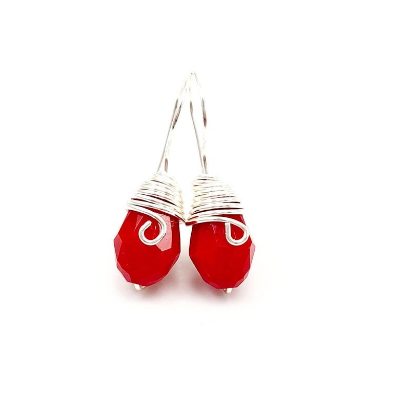 May include: A pair of drop earrings featuring faceted, teardrop-shaped red beads. Each bead is wrapped at the top with silver-colored wire that forms a decorative spiral accent, attached to a simple silver-colored hook for ear suspension.
