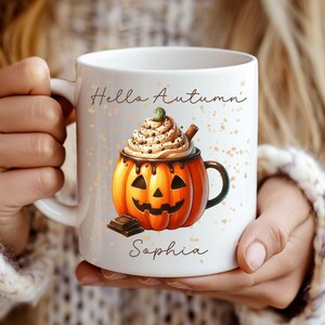 Puede incluir: Taza de cerámica blanca con diseño de calabaza, cubierta de crema batida y palito de canela. La taza tiene las palabras "Hello Autumn" y "Sophia". Decorada con detalles en naranja, marrón y dorado. Tema otoñal.