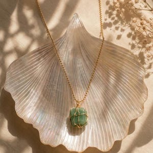 Puede incluir: Un collar de cadena de eslabones esféricos dorados con un colgante de piedra verde de forma cuadrada, encerrado en una delicada jaula de alambre dorado. El colgante cuelga del centro de la fina cadena metálica, mostrando una textura pulida.