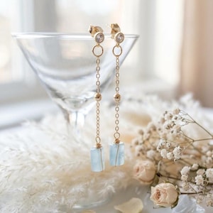Puede incluir: Un par de pendientes colgantes en tono dorado con una pequeña piedra redonda transparente en la parte superior, una cadena delicada y un colgante de piedra rectangular de color azul claro en la base, con acabado metálico pulido.