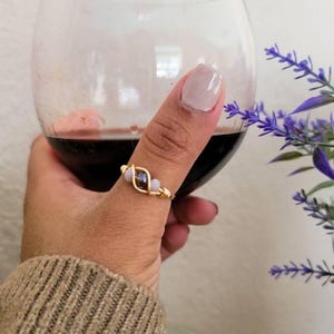 Puede incluir: Un anillo de alambre dorado con una cuenta central oscura flanqueada por dos cuentas más pequeñas de color claro. El anillo está construido con un alambre fino de tono dorado que se enrolla alrededor de las cuentas para asegurarlas en su lugar.