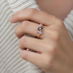 Puede incluir: Un anillo de alambre dorado con una cuenta central de color gris oscuro flanqueada por dos cuentas de color rosa claro. El anillo está en un dedo, mostrando el diseño y el contraste de color. El alambre está intrincadamente tejido, creando una joya única y elegante.