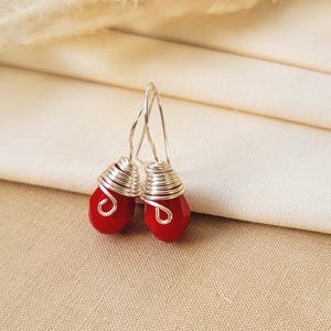May include: A pair of drop earrings featuring faceted red teardrop-shaped beads wrapped in silver-colored wire. The earrings have simple silver-colored wire hooks for attachment, showcasing a clear, vibrant red color and metallic silver wire accents.