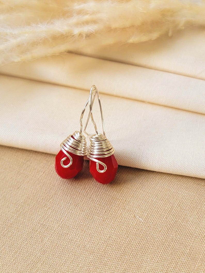 May include: A pair of drop earrings featuring faceted red teardrop-shaped beads wrapped in silver-colored wire. The earrings have simple silver-colored wire hooks for attachment, showcasing a clear, vibrant red color and metallic silver wire accents.