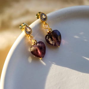 May include: A pair of gold-toned earrings featuring a small, clear, pear-shaped stone at the top, connected by a gold wire to a dangling, heart-shaped, deep purple gemstone. The earrings are displayed on a smooth, white, curved surface.