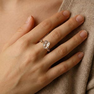 Puede incluir: Un anillo de cuarzo transparente con una piedra en forma de corazón, envuelto en alambre en tonos dorados y plateados. El anillo se muestra en una mano, mostrando el diseño y la artesanía de la joya. El anillo es una pieza de joyería de moda.
