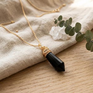 Pode incluir: Um colar de corrente de esferas em tom dourado com um pingente de pedra preta facetada e pontiaguda. O topo da pedra é envolvido por um fio dourado, prendendo-a à corrente. O colar é exibido ao lado de um pequeno grupo de cristais brancos.