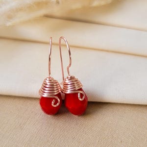 May include: A pair of drop earrings featuring faceted, teardrop-shaped red beads. Each bead is wrapped in a coiled rose gold-colored wire that extends upward into a simple hook-style ear wire, creating a cohesive and metallic design.