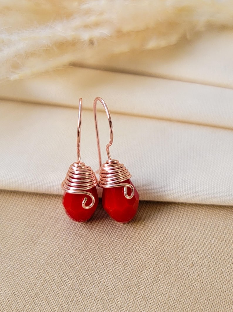 May include: A pair of drop earrings featuring faceted, teardrop-shaped red beads. Each bead is wrapped in a coiled rose gold-colored wire that extends upward into a simple hook-style ear wire, creating a cohesive and metallic design.