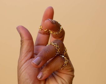 Peut inclure: Un ensemble de bagues dorées présentant un motif répétitif en forme d'étoile. Chaque bague est composée d'une bande continue d'étoiles interconnectées, créant un motif métallique délicat et uniforme qui entoure le doigt avec une finition polie.