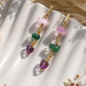Puede incluir: Un par de pendientes colgantes de tono dorado con tres cuentas de piedras preciosas irregulares apiladas en tonos rosa claro, verde y morado oscuro. La cuenta morada inferior tiene forma de corazón y las piedras están unidas por eslabones de alambre dorado.
