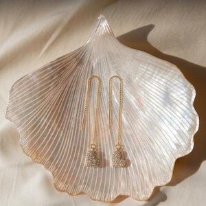 Puede incluir: Un par de pendientes de hilo dorados con cadenas finas que se pasan por la oreja, cada uno terminado en un pequeño dije en forma de corazón incrustado con diminutos cristales transparentes, sobre un plato de concha marina estriada.