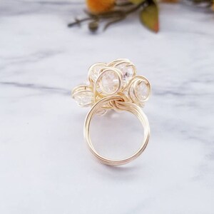 Puede incluir: Un anillo de alambre dorado con un grupo de cristales claros y facetados. Los cristales están dispuestos en un patrón floral o de estrella, con la banda del anillo hecha de alambre enrollado. El anillo está sobre un fondo de mármol.