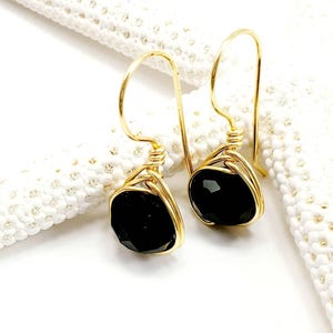 May include: A pair of gold-toned wire-wrapped earrings featuring faceted black stones. Each earring is suspended from a curved gold-toned hook and secured with a decorative wire coil that wraps around the base of the dark, polished gemstone.