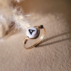 Puede incluir: Un anillo de alambre dorado con una cuenta blanca con la letra "A" en negro. El diseño del anillo incluye un detalle de alambre enrollado alrededor de la cuenta y una banda simple. El anillo está sobre un fondo neutro.