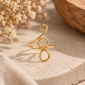 May include: A gold-toned wire ring featuring a continuous design that forms a small circular loop at the top and a figure-eight shape at the center, with the wire wrapping around to create an open band structure with a polished metallic finish.