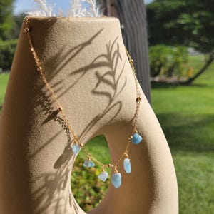 Puede incluir: Un collar de cadena dorada con pequeñas cuentas doradas espaciadas uniformemente y varios dijes colgantes de piedra azul claro, irregulares y pulidos. El collar cuelga sobre un objeto escultórico beige, liso y curvado.