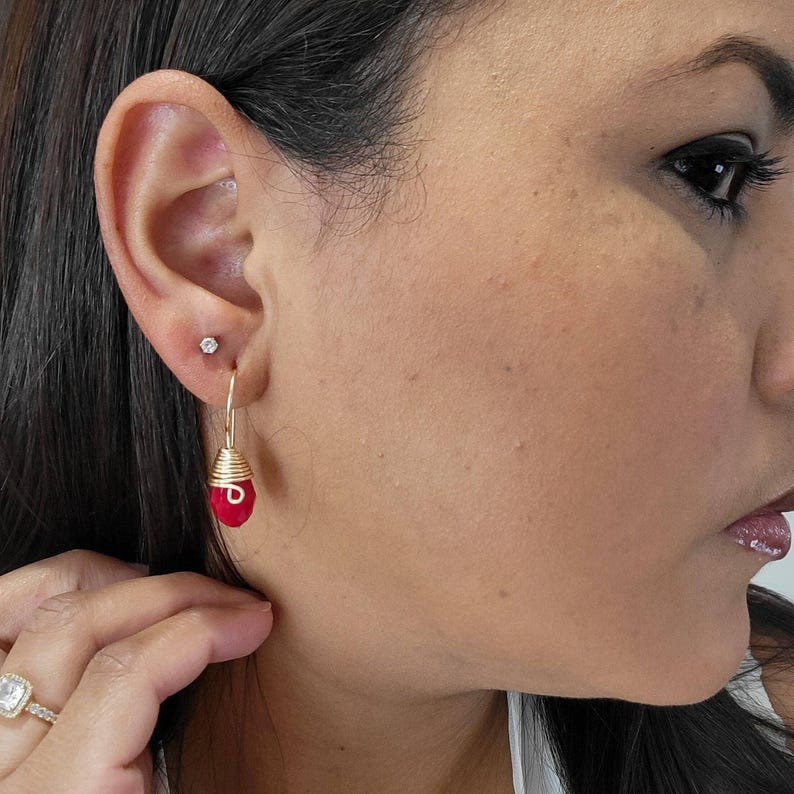 May include: A gold-toned earring featuring a teardrop-shaped red bead wrapped in gold wire, suspended from a hook. A small, clear stud earring is positioned in the upper earlobe, and a gold ring with a clear stone is visible on the finger.
