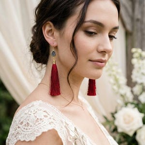 Puede incluir: Un par de pendientes largos colgantes con una piedra gris oscuro en forma de lágrima en la parte superior, conectada a una borla de seda de color rojo intenso y vibrante que cuelga verticalmente. Los pendientes tienen piezas de metal dorado.
