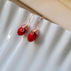 May include: A pair of drop earrings featuring faceted, teardrop-shaped red beads. Each bead is secured with rose gold-colored wire wrapped in a coil design at the top, connected to a matching rose gold-colored curved ear wire hook for suspension.