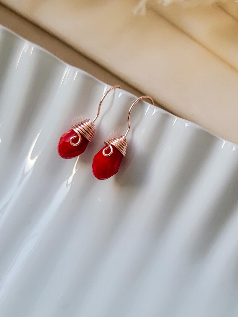 May include: A pair of drop earrings featuring faceted, teardrop-shaped red beads. Each bead is secured with rose gold-colored wire wrapped in a coil design at the top, connected to a matching rose gold-colored curved ear wire hook for suspension.
