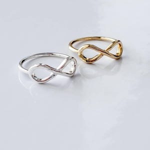 Puede incluir: Dos anillos con un diseño de símbolo de infinito. Un anillo está hecho de metal pulido de tono plateado, mientras que el otro anillo está hecho de metal pulido de tono dorado. Ambos anillos tienen una banda lisa y una estructura de bucle abierto.