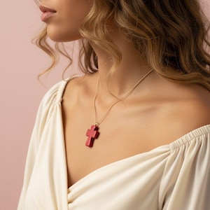 Op de afbeelding: Een goudkleurige ketting met een rode kruis hanger. Het kruis heeft een dieprode kleur en een getextureerd oppervlak. De ketting wordt gedragen over een crèmekleurige top.