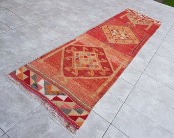 May include: A long, rectangular, hand-woven rug with a vibrant red and orange color scheme. The rug features geometric patterns in shades of orange, tan, and red. The edges are finished with a fringe. This rug is suitable for a hallway or entryway.