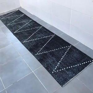 May include: A long, rectangular black rug with a geometric pattern of white dotted lines. The rug is placed on a tiled floor, and the design creates a zig-zag effect across the length of the rug. The rug is likely made of a soft material.