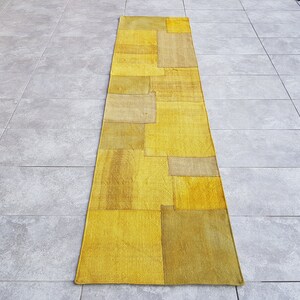 Könnte beinhalten: Ein langer, rechteckiger gelber und beiger Patchwork-Teppich mit geometrischem Muster. Der Teppich besteht aus verschiedenen Stoffstücken, die zusammengenäht wurden.