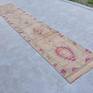 May include: A long, rectangular rug with a beige background and intricate patterns in pink, orange, and brown. The rug features geometric designs and a border with pink triangles. Ideal for a hallway or entryway.