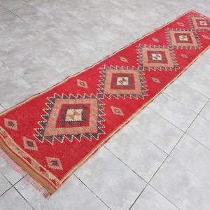 May include: A red and beige patterned runner rug with a geometric design. The rug has a fringe border.