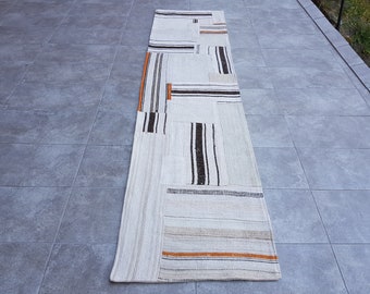 May include: A white and beige runner rug with black and orange stripes. The rug is made of woven fabric and has a patchwork design.