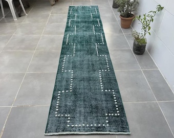 May include: A long, rectangular, dark teal rug with a geometric pattern of white dots. The rug is placed on a tiled floor and appears to be made of a woven material. The design features a series of connected shapes.