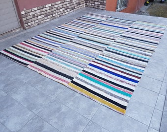 May include: A colorful striped rug made from recycled materials. The rug is woven with a variety of colors, including black, white, blue, green, and yellow. The rug is laid out on a gray tiled floor.