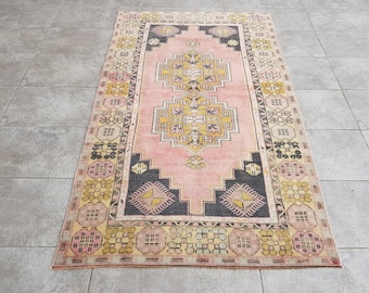 May include: A rectangular rug with a pink field and geometric patterns. The central design features a yellow and black diamond pattern. The border has a repeating pattern of yellow and black shapes. The rug is likely made of wool or a similar material.