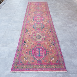 May include: A long, pink and brown patterned rug with a geometric design. The rug has a fringe border and is laid out on a gray tiled floor.