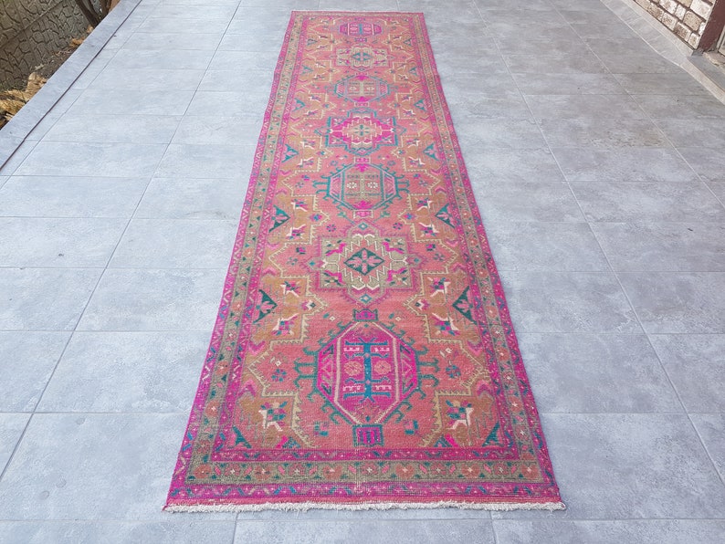 May include: A long, pink and brown patterned rug with a geometric design. The rug has a fringe border and is laid out on a gray tiled floor.