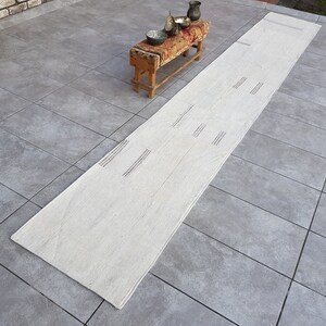 May include: A long, off-white runner rug with a subtle woven pattern. The rug is laid out on a gray tiled floor.