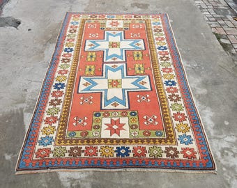 May include: A rectangular area rug featuring a vibrant orange field with two large central light blue and white cross motifs. The border displays a repeating pattern of small geometric shapes in red, blue, yellow, and green, with a fringed edge.