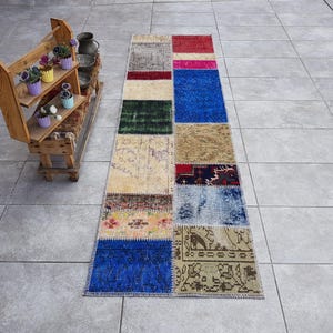 Op de afbeelding: Een rechthoekig patchwork vloerkleed met een verscheidenheid aan kleuren en patronen. Het vloerkleed heeft vierkanten in rood, roze, blauw, groen en beige. Het vloerkleed ligt op een grijze betegelde ondergrond.