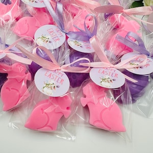 May include: An assortment of Easter-themed party favors. The favors include pink and purple bunny-shaped soaps, wrapped in clear cellophane bags and tied with pink and purple ribbons. Each favor has a circular tag that reads "Happy Easter".