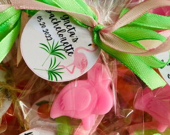 May include: Pink flamingo-shaped party favours, individually wrapped and tied with green and pink ribbons. Each favour has a tag that reads "Greta's Bachelorette 05.20.2022" with a flamingo illustration. The favours are ideal for a bachelorette party.